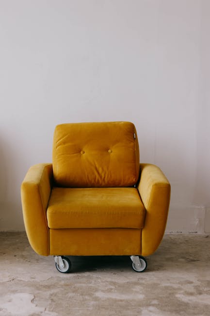 A person using a handheld vacuum cleaner to deep clean a decorative cushion with a yellow, white, and grey geometric pattern on a dark fabric sofa in a well-lit room. The individual is wearing a dark 