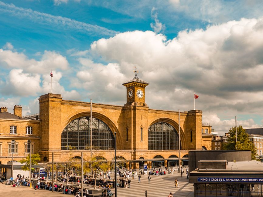 A large, historic train station building made of yellowish stone with arched glass windows and a central clock tower topped with a weather vane. The station is bustling with people outside, some sitti